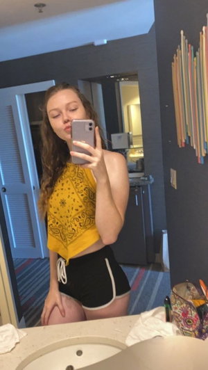 Woman in yellow crop top and black shorts taking mirror selfie in bathroom.