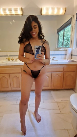 Woman in black mesh lingerie taking mirror selfie in bathroom.