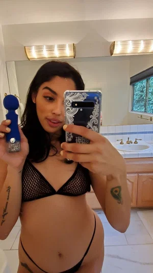 Woman in black mesh lingerie holding a toy, taking mirror selfie in bathroom.