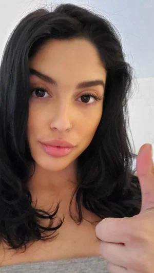 Woman with dark hair and gray top giving thumbs up indoors.