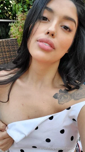 Woman with dark hair and chest tattoo wearing white polka-dot top outdoors.