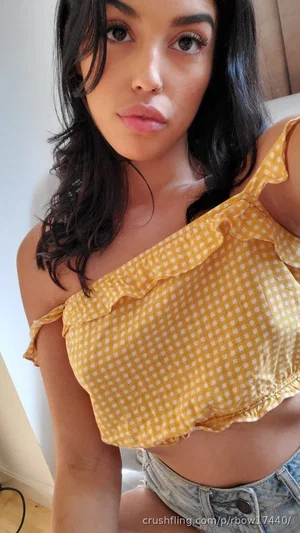 Woman with dark hair in yellow gingham top and denim shorts, sitting indoors.