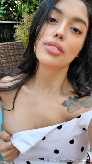 Woman with dark hair and rose tattoo on shoulder, wearing white polka-dot top, outdoors.