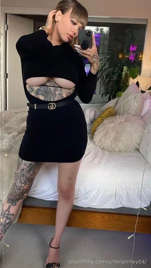 Woman in black cutout dress with tattoos, posing in bedroom selfie.