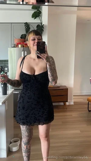 Woman with tattoos in black dress taking mirror selfie in kitchen.