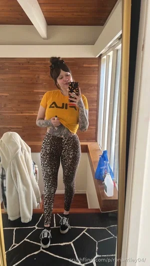 Woman with tattoos wears yellow shirt and leopard leggings, standing in mirror selfie in indoor