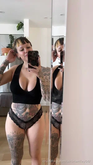 Woman with tattoos in black top and lace underwear takes mirror selfie in bedroom.