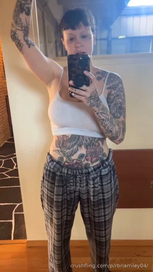 Woman with tattoos in white crop top and plaid pants taking mirror selfie indoors.