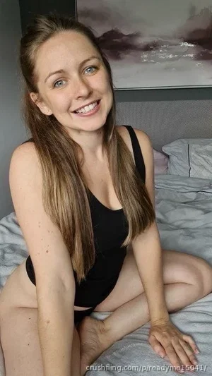 Woman with long brown hair in black bodysuit sitting on bed, smiling at camera.