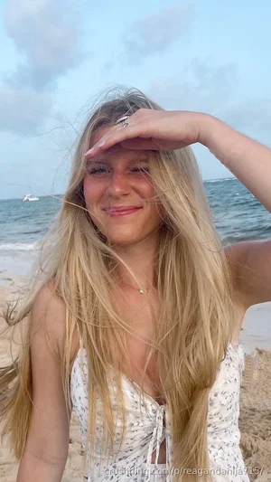 Blonde woman in floral top shielding eyes at beach, smiling.