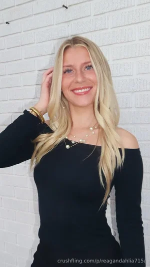 Blonde woman in black off-shoulder top, smiling, standing by white brick wall.