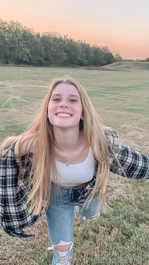 Woman with long blonde hair in a white top and plaid shirt, standing in a grassy field at sunset.