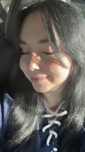 Woman with long dark hair wearing a dark blue top with white lace-up front, sitting in a car.