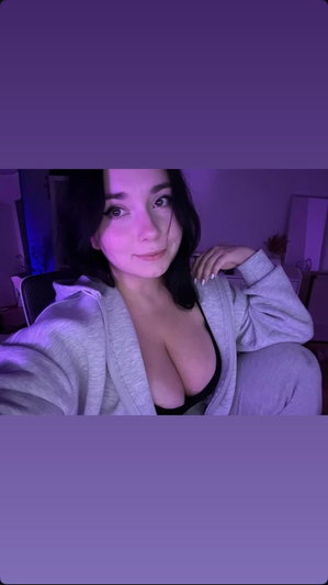 Woman in gray hoodie and black top, sitting indoors under purple lighting.