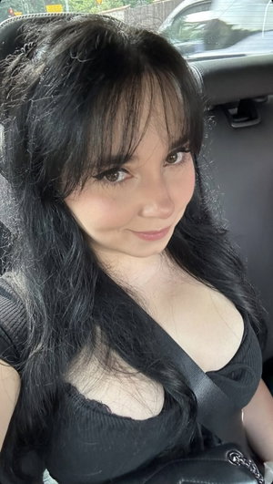 Woman with long black hair in black top sitting in car, smiling at camera.