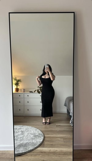 Curvy woman in black dress in bedroom mirror selfie.