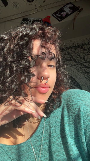 Woman with curly hair and nose ring wearing teal top indoors.