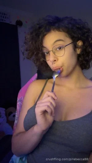 Woman with curly hair and glasses in a gray tank top holding a spoon indoors.