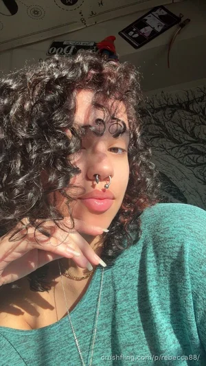 Woman with curly hair and nose rings wearing a green top indoors.