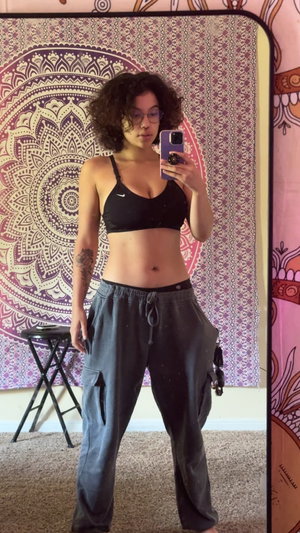 Woman with curly hair in black sports bra and gray pants taking mirror selfie indoors.