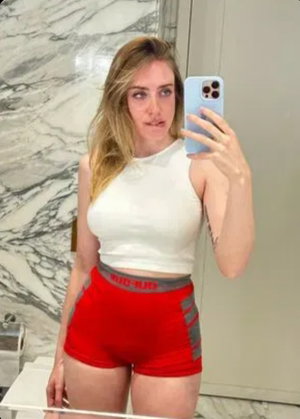 Woman with blonde hair in white crop top and red shorts taking mirror selfie in marble bathroom.