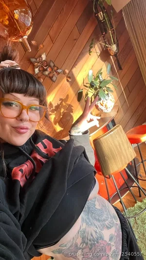 Woman with glasses and tattoos in black hoodie, indoors with plants and wooden decor.