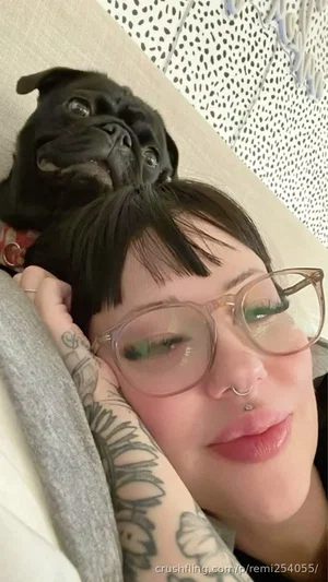 Woman with short black hair, glasses, and arm tattoos lying on bed with black pug on head.