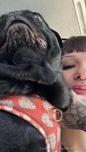 Woman with tattoos and nose ring posing with black pug wearing red harness.