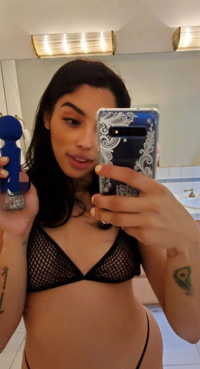 Woman with dark hair in mesh bra taking bathroom mirror selfie.
