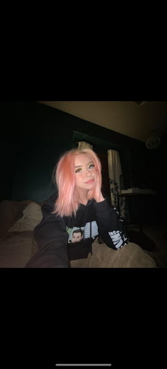 Woman with pink hair in black sweatshirt sitting on bed in dimly lit room.