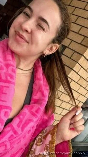 Woman with long brown hair wearing pink patterned robe over dark top, smiling in front of brick