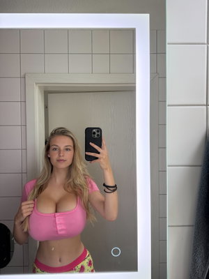 Blonde woman in pink crop top and patterned shorts taking mirror selfie in tiled room.