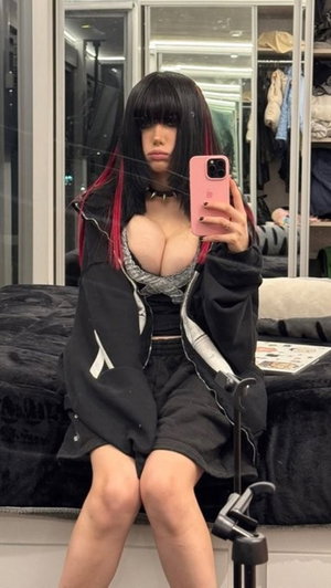 Woman with black and pink hair in a black jacket and crop top, sitting on bed in bedroom, taking