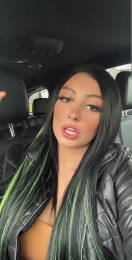 Woman with long black and green hair in a black jacket, sitting in a car.