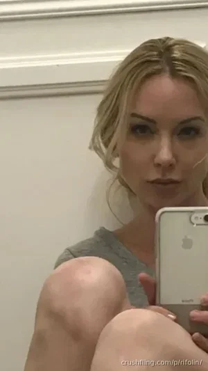 Blonde woman in gray top taking mirror selfie, legs visible, indoor setting.