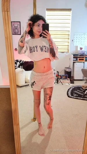 Woman with tattoos in a crop top and shorts takes mirror selfie in bedroom.
