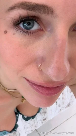 Close-up of a woman with blue eyes, freckles, and a nose ring, wearing a patterned top indoors.
