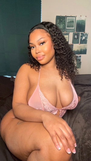 Woman with curly hair in pink lace lingerie sitting on bed indoors.