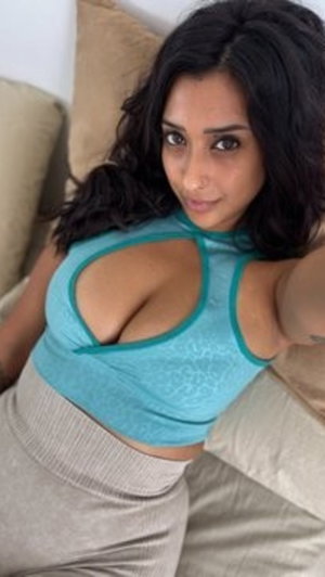 Woman with dark hair in blue crop top and beige skirt, posing on couch indoors.