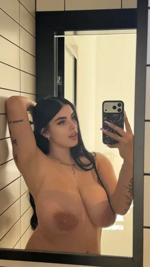 Curvy woman with dark hair taking bathroom mirror selfie topless.