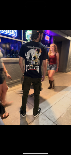 Man in black t-shirt and cap standing on city sidewalk at night.