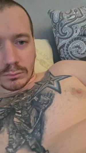Man with tattoos on chest and shoulder lying in bed, indoor setting.