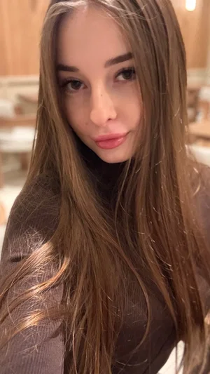 Woman with long brown hair wearing a dark turtleneck in a softly lit indoor setting.