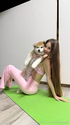 Woman with long hair in pink workout outfit posing with a puppy on a green mat indoors.