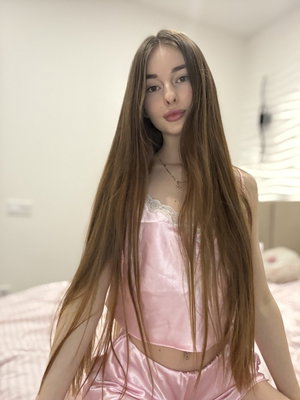Woman with long brown hair wearing pink satin pajamas in bedroom.