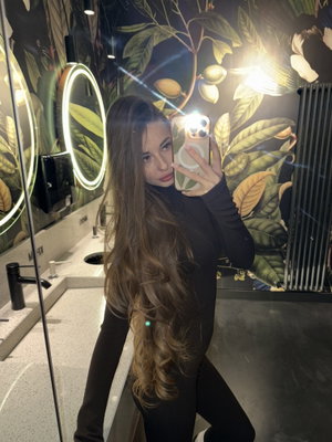 Woman with long hair in black outfit takes mirror selfie in bathroom with tropical wallpaper.