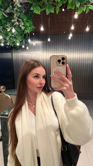 Woman with long brown hair in white fuzzy sweater and scarf, taking mirror selfie in indoor setting
