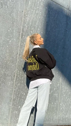 Blonde woman in black jacket and light jeans standing by metal wall outdoors.