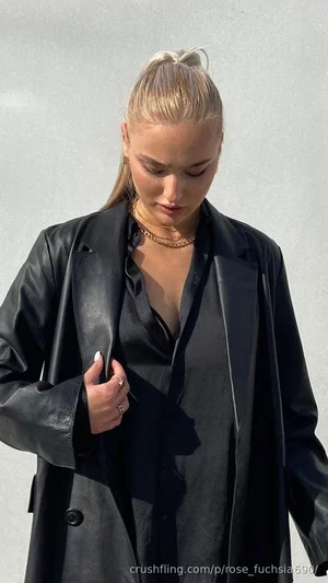 Blonde woman in black leather coat and silk top, standing against a white wall.