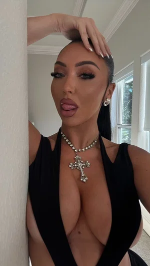 Woman in black plunging top with cross necklace, posing indoors near wall.